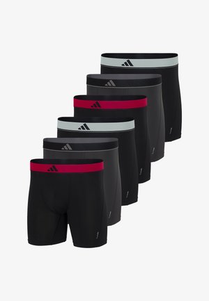 Athletischer Mann steht barfuß, trägt schwarze Boxershorts mit rotem Bund und Adidas-Logo, schaut leicht zur Seite.