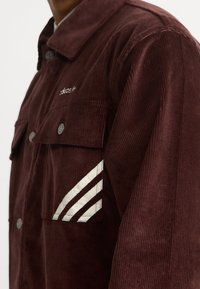 Burgundy kordjacka med silverdetaljer, två bröstfickor och vit tre-randig design på vänster ärm.
