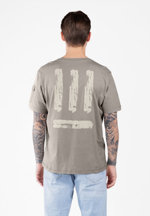 Hombre con tatuajes en los brazos que lleva una camiseta gris claro con tres diseños de trazos de pincel verticales y uno horizontal en color beige en la parte de atrás.