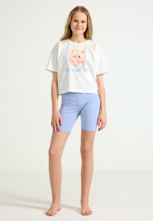 Teini-ikäinen tyttö seisoo paljain jaloin valkoisessa lyhyessä T-paidassa, jossa on kissakuvio, ja vaaleansinisissä pyöräilyshortseissa valkoista taustaa vasten.