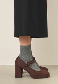 Pantofi cu toc din suede maro, cu vârful rotund și o curea cu cataramă argintie, purtați cu șosete gri cu dungi, pe un fundal neutru.
