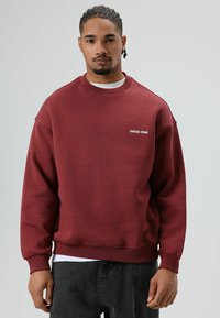 Sweatshirt bordeaux en tissu doux avec un col rond et des épaules tombantes. Présente un texte blanc "RAÍCES VIVAS" sur la poitrine.