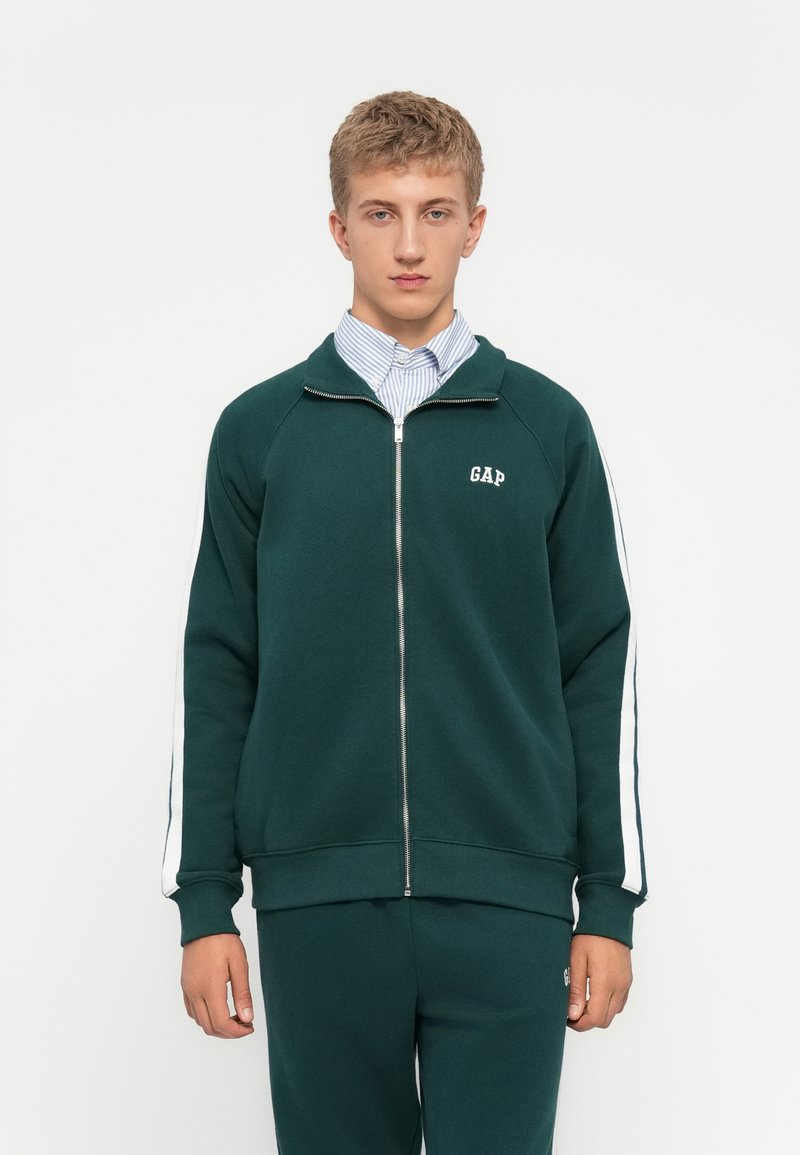 Green zip-up sweatshirt with white side stripes, made of soft fabric. Features a small "GAP" logo on the chest. Paired with matching pants.
