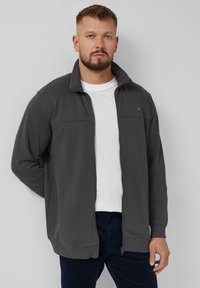 Giacca grigia con zip e colletto alto, dotata di due tasche frontali, realizzata in morbido tessuto. Indossata sopra una t-shirt bianca con pantaloni scuri.