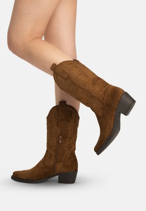 Ben iført mellemstore brun ruskinds cowboystøvler med dekorative syninger, sidelås og blokhæle mod en hvid baggrund.