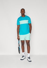 Türkises Sportshirt mit einem hellblauen Streifen, kombiniert mit mintgrünen Shorts. Trägt weiße und marineblaue Tennisschuhe und hält einen Tennisschläger.