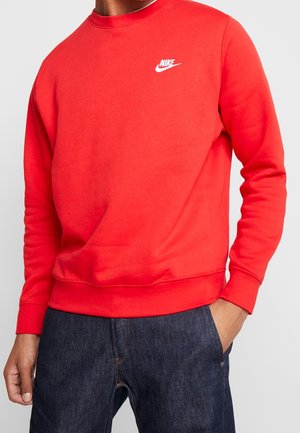 Homme portant un sweat à capuche Nike rouge vif et un jean bleu foncé, la main dans la poche, sur un fond clair.