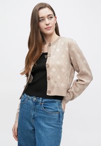 Μπεζ ζακέτα με ανοιχτόχρωμα λουλουδάτα σχέδια, στρογγυλή λαιμόκοψη και κουμπιά στο μπροστινό μέρος. Φοριέται πάνω από μαύρο αμάνικο τοπ, σε συνδυασμό με μπλε τζιν. Ομαλή υφή.