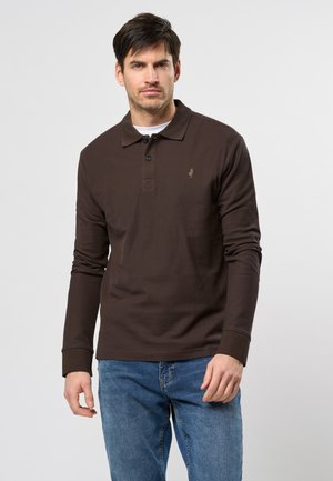 Moški nosi temno rjavo dolgo majico polo nad belo podmajico, v kombinaciji z modrimi džinsom, stoji ob enotni ozadju.