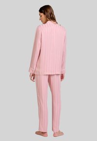 Mujer de pie descalza, de espaldas, vistiendo pijamas de rayas rosa claro con mangas largas y pantalones a juego, frente a un fondo liso.