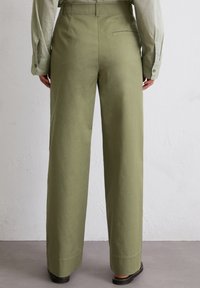 Wide-leg trousers in olive green cotton, featuring a back pocket, smooth texture, and straight hem. Worn with a light-colored shirt.