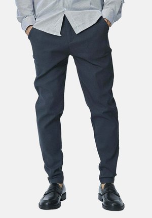Homme portant un pantalon fuselé bleu marine foncé, des mocassins noirs, et une chemise gris clair boutonnée rentrée, les mains dans les poches.
