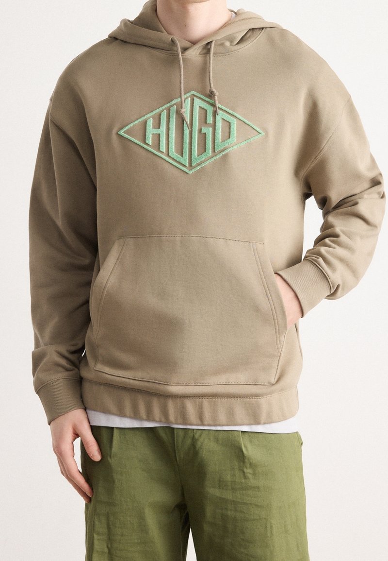 Felpa khaki con cappuccio e tasca frontale. Presenta un logo a forma di diamante di colore teal "HUGO" al centro. Sono visibili dettagli cuciti.