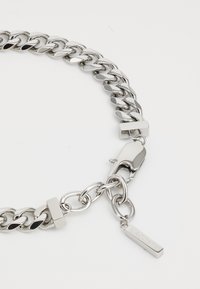 Bracelet en chaîne argentée avec des maillons entrelacés, un fermoir mousqueton et une étiquette rectangulaire suspendue embossée du nom de la marque.