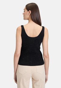 Débardeur noir à côtes avec un dos plongeant, présentant un tissu texturé et des détails de léger éclat éparpillés. Associé à un pantalon beige clair.