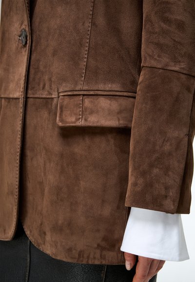 Barna velúr dzseki zsebfedéllel az ujjon, fehér ing mandzsettája és fekete nadrág fölött, részben látható személy karján.