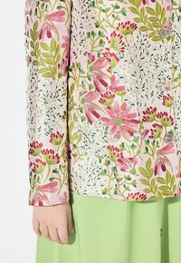 Blazer floreale in tessuto rosa chiaro con motivi botanici verdi e rossi, due bottoni e una tasca. Indossato sopra una gonna verde.
