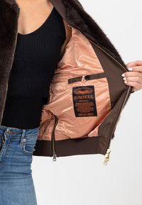 Bruine bomberjack met een zachte nepbontkraag, satijnen perzikkleurige voering, gouden ritssluiting en oranje labeldetails. Klassiek ontwerp en kenmerken.