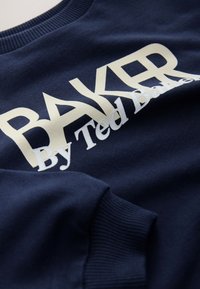 Marineblauwe sweatshirt met geribde halslijn, voorzien van gedurfde beige gedrukte tekst: "BAKER" en "By Ted Baker." Glad stoffenstructuur.