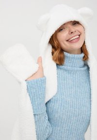 Biała koc z kapturem i uszami misia. Miękka, puszysta tekstura. Jasnoniebieski sweter z golfem o prążkowanym wzorze pod spodem.