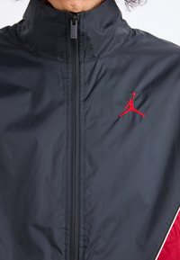 Black windbreaker jacket with high collar and full zip front. Features a red embroidered logo on the chest and red accents on the sides.