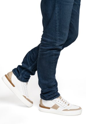 Beine in dunkelblauer Jeans und weißen Sneakers mit beigen Akzenten, ein Fuß angehoben, als würde er gehen oder einen Schritt nach vorne machen.