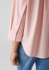 Hellrosa Langarmshirt mit Knopfmanschetten, weichem Stoff und abgerundetem Saum, getragen über blauen Jeans.