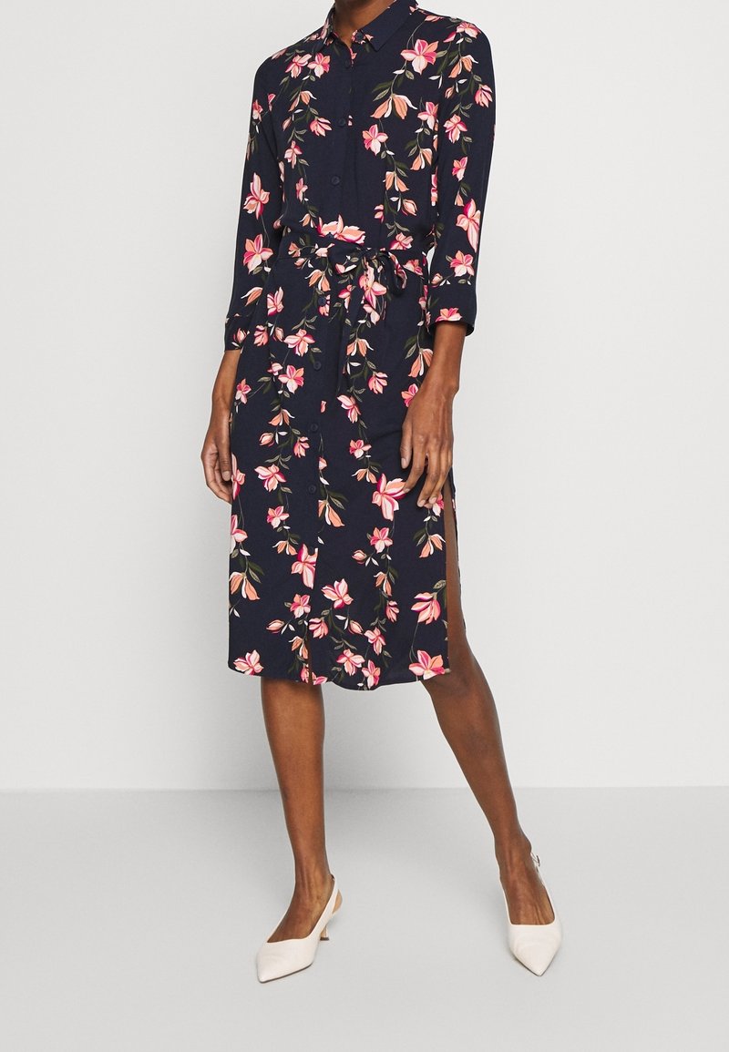Femme portant une robe midi bleu marine à fleurs, manches longues, ceinture à la taille, boutons sur le devant, fente latérale et talons blancs à bouts pointus.