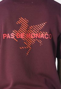 Burgundy paita, jossa on punaraitainen hevostyynykuva ja teksti "PAS DE MONACO" rinnassa.