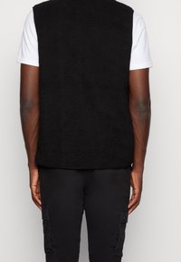 Osoba v černé texturované bezrukávové vestě přes bílou košili s krátkým rukávem a černých cargo kalhotách, zobrazená zezadu.