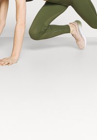 Oliwkowe legginsy charakteryzują się teksturowanym wzorem oraz wycięciami wzdłuż dolnej części nogawek, w połączeniu z jasnoróżowymi butami sportowymi.