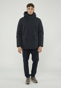 Giacca puffer blu navy con cappuccio, dotata di zip frontale e chiusura a scatto, due tasche frontali e un tessuto liscio e resistente. Abbinata a pantaloni scuri.