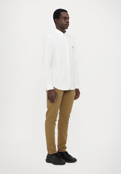 Vit skjorta med knappar, bruna byxor och svarta sneakers. Slim fit, lång ärm, subtilt varumärke på skjortan. Slät tyg och enfärgade.
