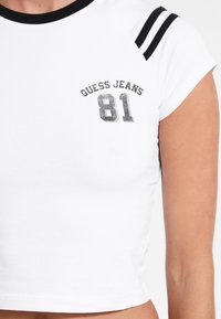 Balta trumpa marškinėliai su juodais akcentais. Turi apvalų kaklą ir trumpas rankoves. Priekyje užrašyta "GUESS JEANS" ir "81" sidabriniu spaudu.