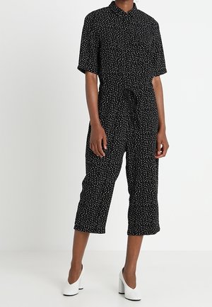 Vrouw in een zwart cropped jumpsuit met witte spikkels, korte mouwen en witte blokhakken, staand tegen een effen achtergrond.