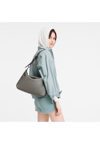 Graue Lederhandtasche mit geschwungener Form und verstellbarem schwarzen Tragegurt. Das Modell trägt ein hellblaues Satinhemd und gestreifte grüne Shorts.