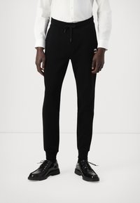 Schwarze Jogginghosen mit elastischem Bund und Kordelzug. Enganliegende Bündchen und kleines Logodetail an der Seite. Kombiniert mit schwarzen Schnürschuhen.