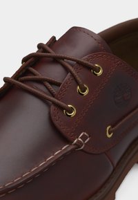 Καφέ δερμάτινο παπούτσι τύπου boat με στρογγυλά κορδόνια, χρυσά μεταλλικά τρύπες, λεπτομερείς ραφές και ανάγλυφο λογότυπο δέντρου στο πλάι.