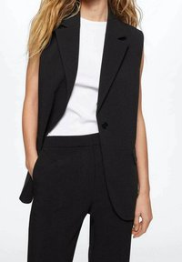 Blazer sans manches noir en tissu lisse, avec une fermeture à un bouton, des revers en pointe et des poches latérales, associé à une chemise blanche.