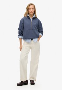 Un sweat à capuche bleu marine avec une fermeture éclair à mi-hauteur, associé à une chemise rayée bleu clair et blanche, un pantalon large crème et des mocassins foncés.