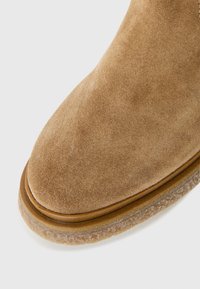 Wildleder-Ankle-Boots in Beige mit rundem Zehenbereich, ausgestattet mit einem gesteppten Lederbesatz und einer strukturierten Gummisohle. Glatte Oberfläche, keine sichtbaren Muster.
