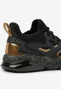 Czarne buty sportowe z siateczkowym i syntetycznym wierzchem, metaliczne brązowe akcenty, nakrapiana szara podeszwa oraz tylny uchwyt ułatwiający zakładanie.