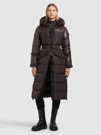 Lang mørk puffet frakke med quiltet tekstur, hætte, indsnævret talje og forlommer. Kombineret med sorte leggings og chunky sorte støvler.