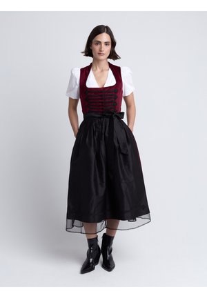 Frau trägt ein burgunderfarbenes Korsett-Top mit schwarzen Knöpfen, weiße Bluse mit Puffärmeln, schwarzen Midirock mit Schleife und schwarze Stiefeletten.
