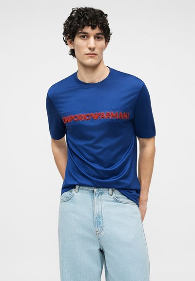 Άνδρας με σγουρά σκούρα μαλλιά που φοράει μπλε μπλούζα Emporio Armani με κόκκινο λογότυπο και ανοιχτόχρωμο τζιν, στέκεται μπροστά σε απλό φόντο.