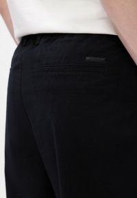 Pantaloni negri din bumbac, cu un buzunar la spate ce prezintă un eticheta subtilă a brandului. Materialul are o textură netedă și o croială ajustată.