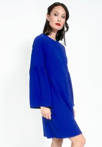 Rochie albastră cu mâneci scurte, guler rotund și mâneci în formă de clopot. Țesătură netedă cu o ușoară strălucire, tăiată până la genunchi, cu detalii minime.