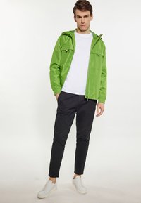 Jachetă impermeabilă verde cu fermoar, buzunare cargo și glugă ajustabilă, asociată cu pantaloni închiși la culoare și adidași albi, pe un fundal neutru.