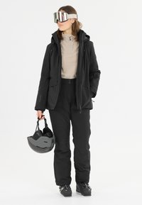 Skidkläder bestående av en svart vattentät jacka och byxor, i kombination med en beige topp, svart hjälm och spegelgoggles.