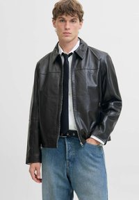 Schwarze Lederjacke mit Kragen, Reißverschluss und zwei Seitentaschen. Über einem weißen Hemd und einer Krawatte getragen, kombiniert mit hellblauer Jeans.